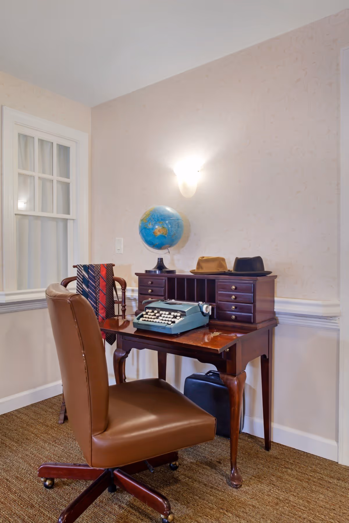 A vintage-style office corner with a wooden desk holding a blue typewriter, a globe, and two hats. A brown leather office chair is positioned in front of the desk. A wooden rack with several ties is placed beside the desk. The room has beige wallpaper with a subtle pattern and a wall-mounted light fixture above the desk.