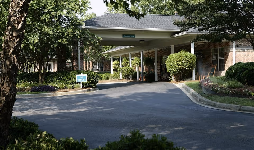 Entrance driveway of a senior living facility with a covered drop-off area, surrounded by trees, bushes, and a brick building. A sign reads 'NO TRUCKS' and there are rocking chairs on the porch.
