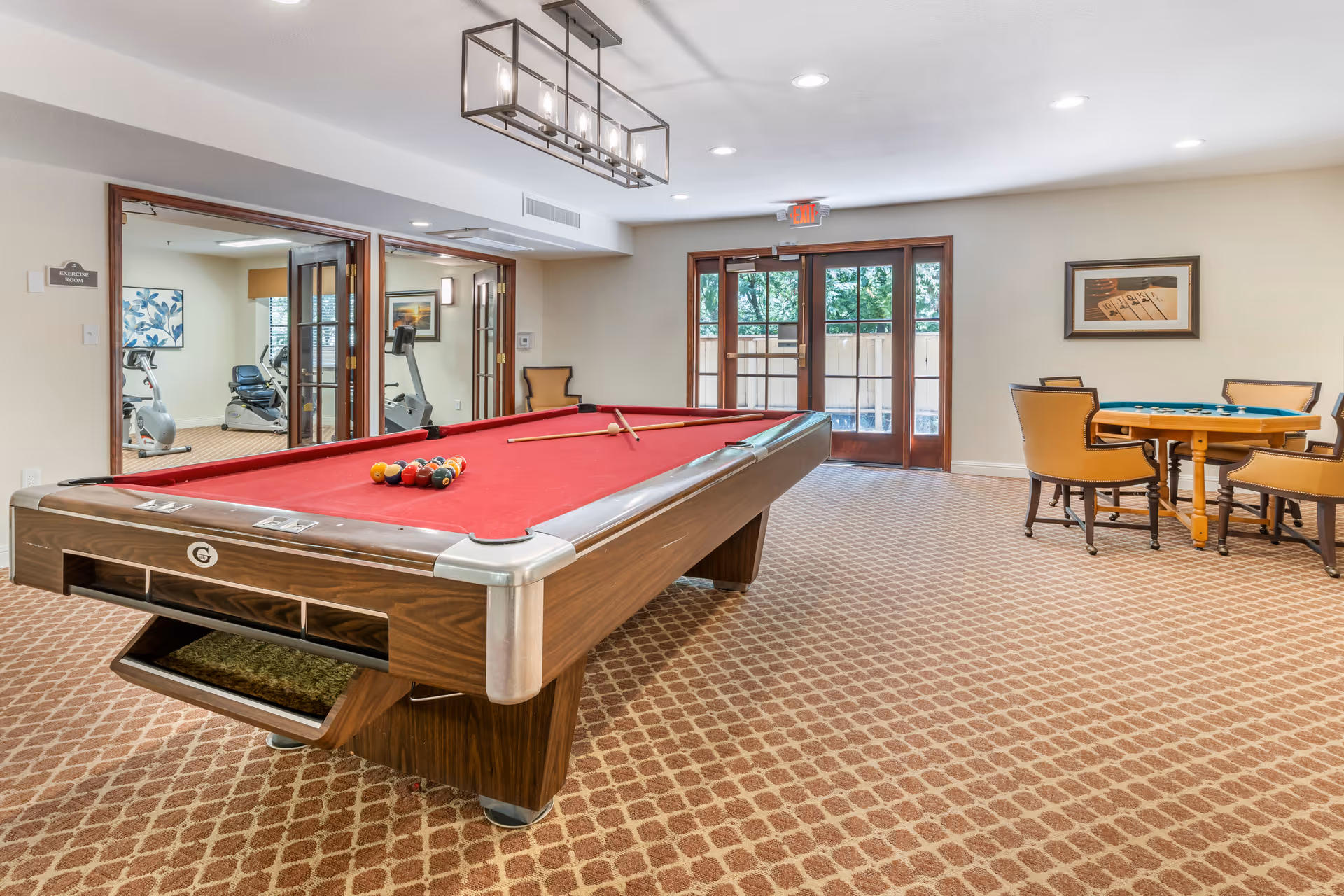 A recreational room with a red felt pool table in the center, a card table with four chairs to the right, and a view into an exercise room with fitness equipment through glass doors on the left. The room has patterned carpet, beige walls, and a modern rectangular ceiling light fixture. Double glass doors at the back lead outside, letting in natural light.