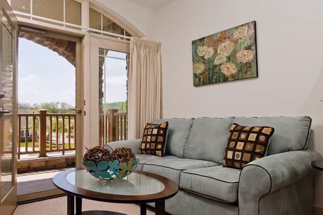 A cozy living room with a light green upholstered sofa adorned with two patterned throw pillows. In front of the sofa is a round glass-top coffee table with a decorative bowl containing natural elements. Behind the sofa, a floral painting hangs on the white wall. To the left, glass doors open to a balcony with wooden railings and a view of greenery outside.