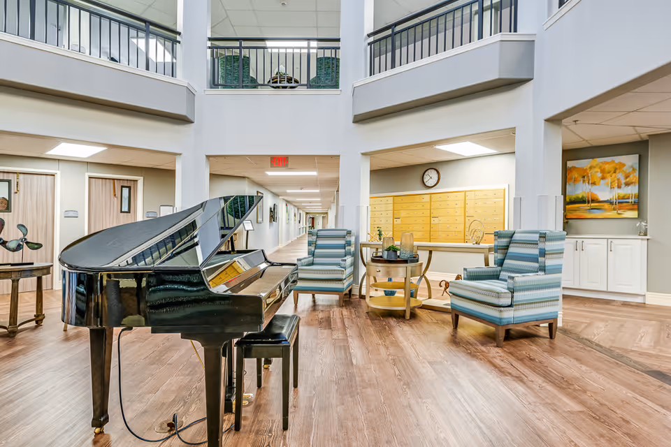 Interior view of a senior living facility common area featuring a black grand piano with a matching bench, two striped armchairs, a round wooden table with decorative items, and a wall of mailboxes with a clock above. The space has wood flooring, light-colored walls, and a high ceiling with a balcony railing visible above.