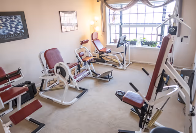 Small exercise room with multiple red-and-white weight and leg-press machines, carpeted floor, and a large window letting in natural light.