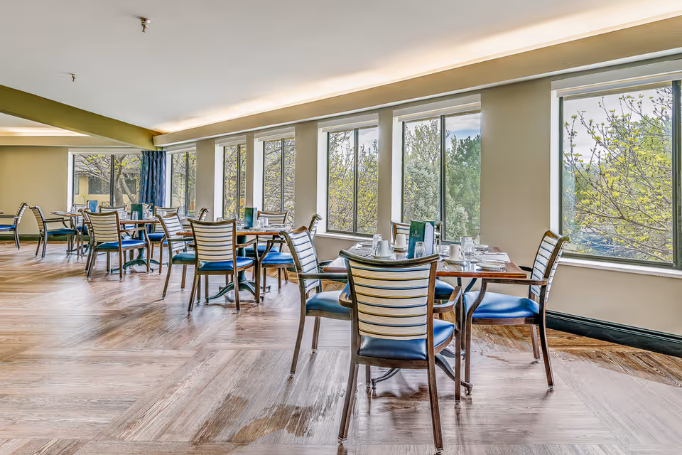 A bright dining room with multiple tables and chairs arranged neatly. Each table is set with white cups, glasses, and menus. Large windows line the wall, allowing natural light to fill the room and providing a view of trees outside.