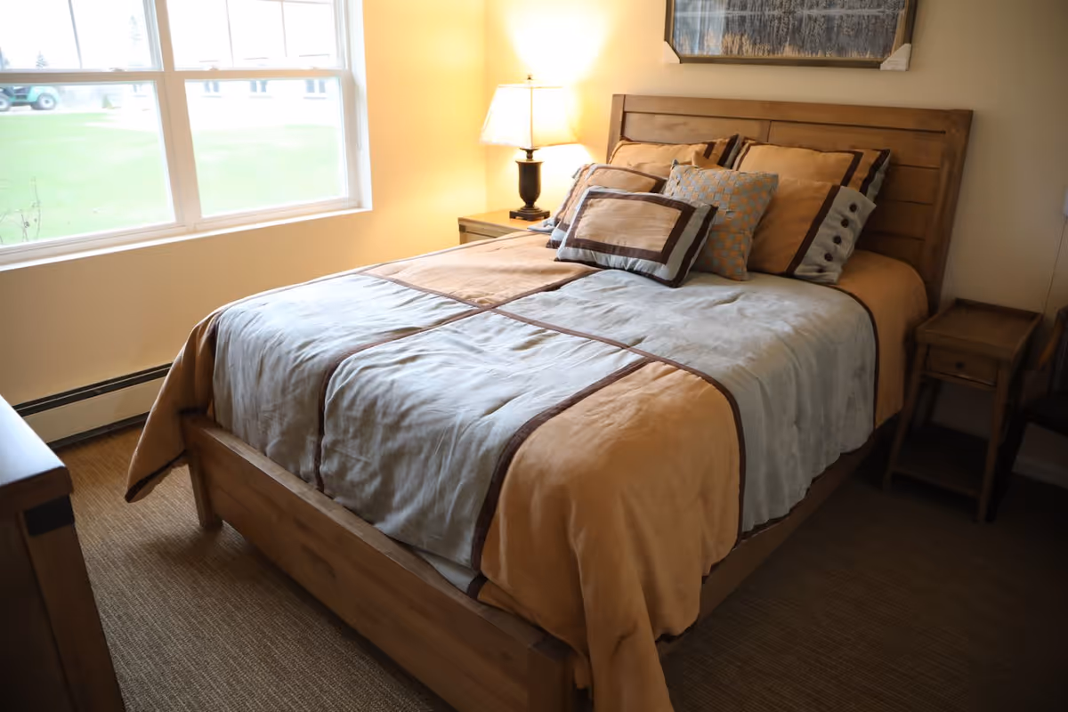 A neatly made bed with a wooden frame and headboard in a bedroom. The bed has a beige and light blue comforter with multiple decorative pillows. There is a nightstand with a lamp on the right side of the bed and a window letting in natural light on the left side.