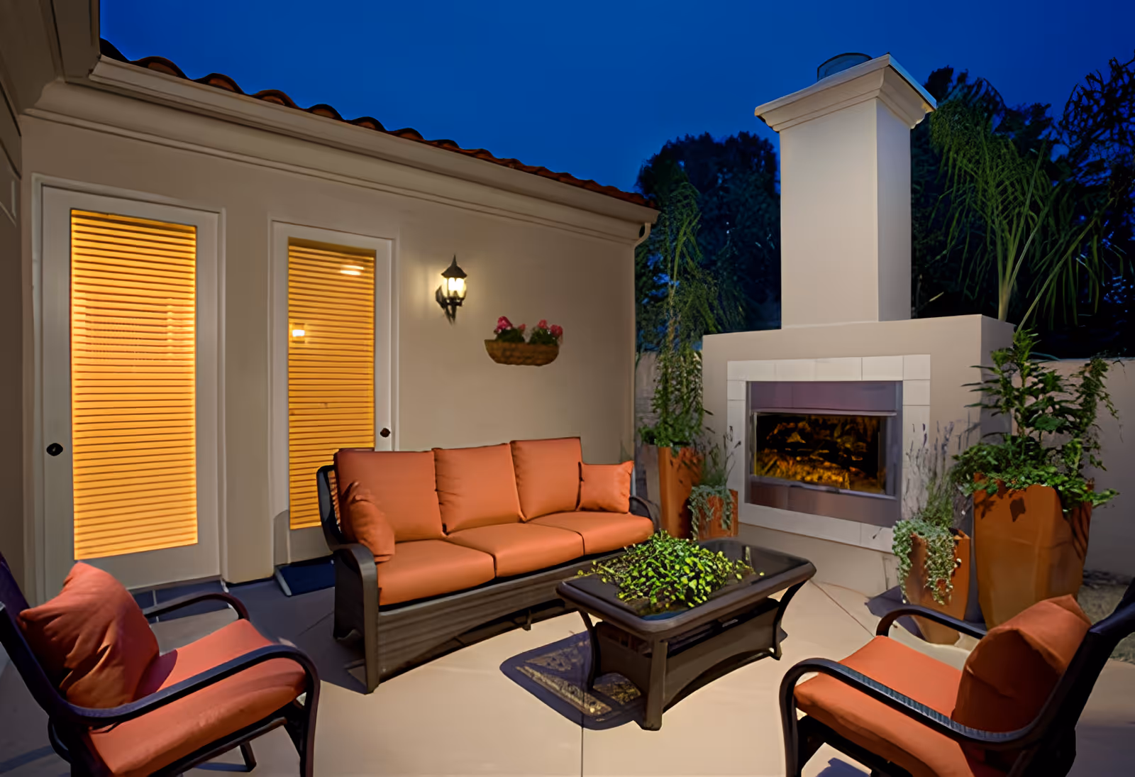 Evening outdoor patio with orange-cushioned seating arranged around a coffee table and a built-in fireplace.