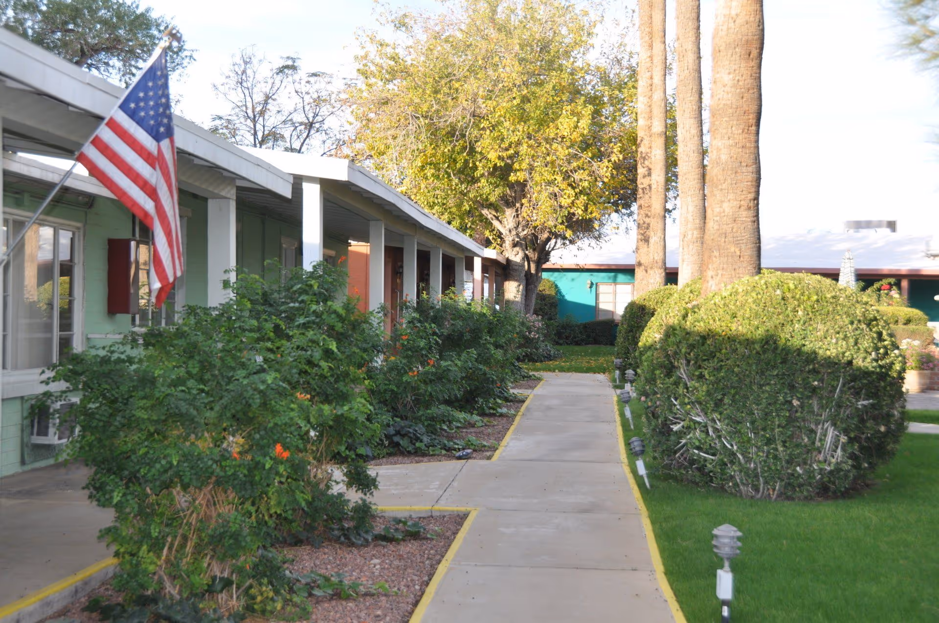 A walkway lined with bushes and trees runs between single-story buildings with covered porches. An American flag is mounted on one of the buildings. The area is well-maintained with green grass and trimmed shrubs under a clear sky.