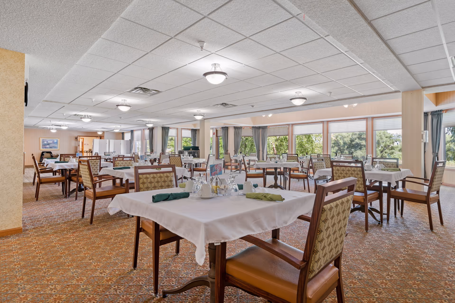 A spacious dining room with multiple tables covered in white tablecloths, each set with glasses, napkins, and condiments. The room has large windows with curtains allowing natural light and a view of greenery outside. The carpet has a patterned design, and the ceiling features multiple light fixtures.