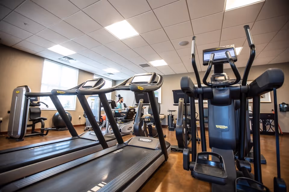 Indoor fitness room with treadmills, elliptical machines, and other exercise equipment. A person is using a recumbent bike in the background near a window with natural light.