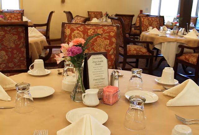 A dining room table set with beige tablecloth, white plates, upside-down glasses, folded napkins, silverware, a small vase with pink and purple flowers, a menu card, and a container with pink sweetener packets. In the background, there are more tables and chairs with similar settings and patterned upholstery.