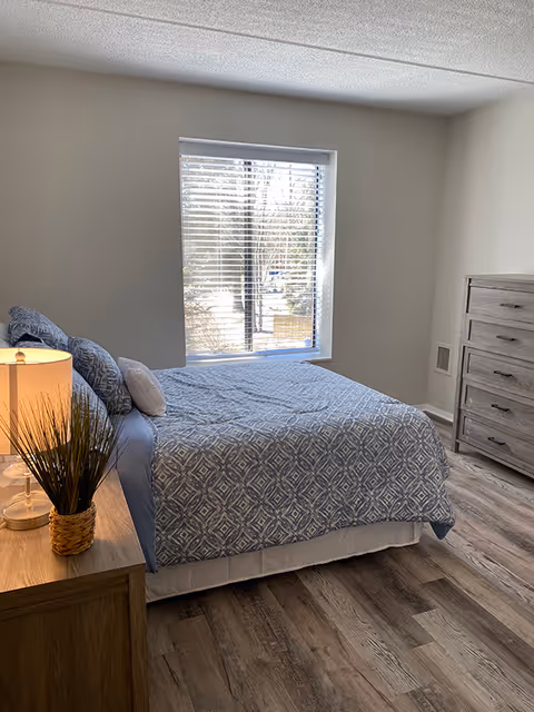 A neatly made bedroom with a patterned bed by a window, a wooden nightstand with lamp and plant, and a tall dresser on wood flooring.