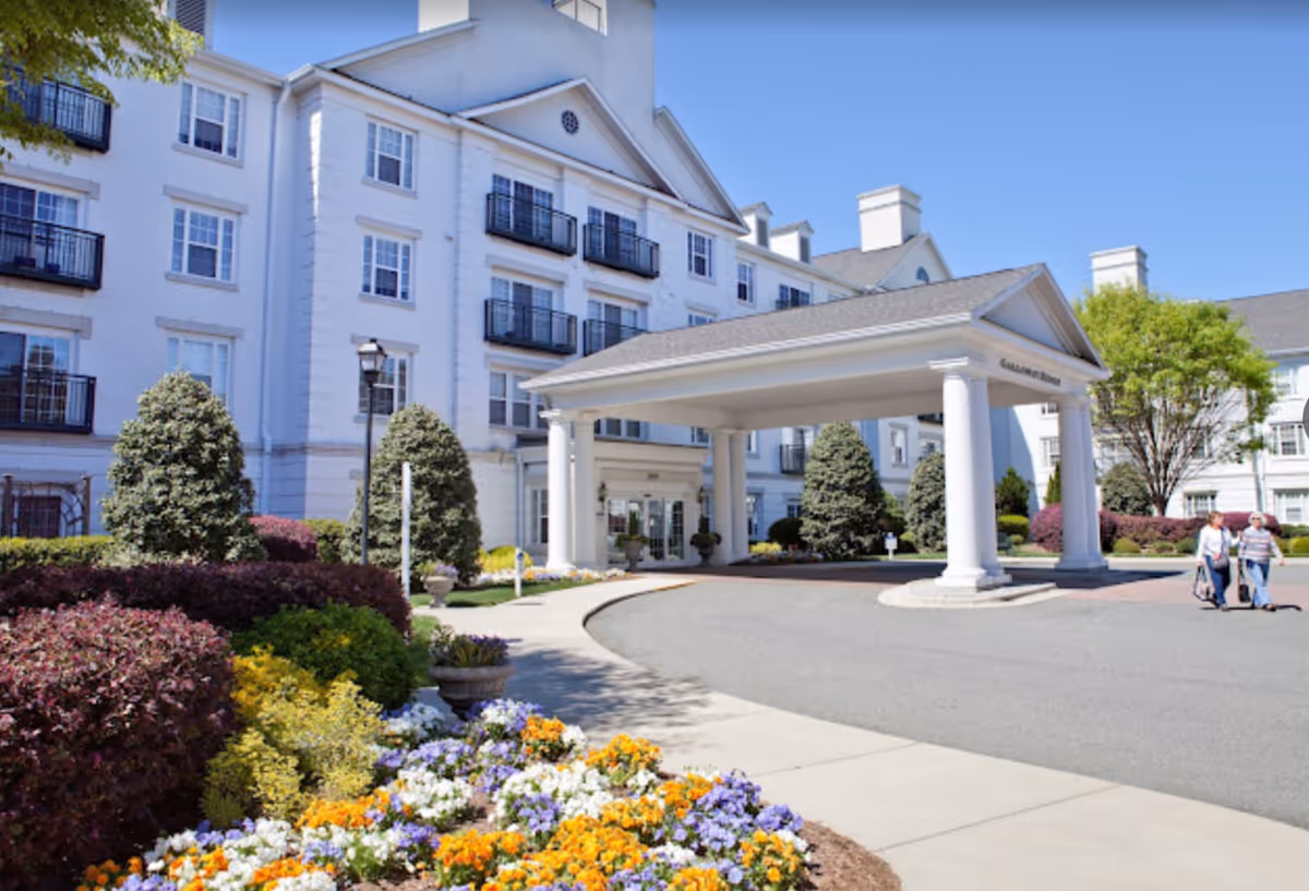 Exterior view of Galloway Ridge at Fearrington, a large white multi-story building with black balconies. The entrance features a covered driveway supported by white columns. There are well-maintained flower beds with colorful flowers and shrubs along the sidewalk, and two people walking near the entrance.