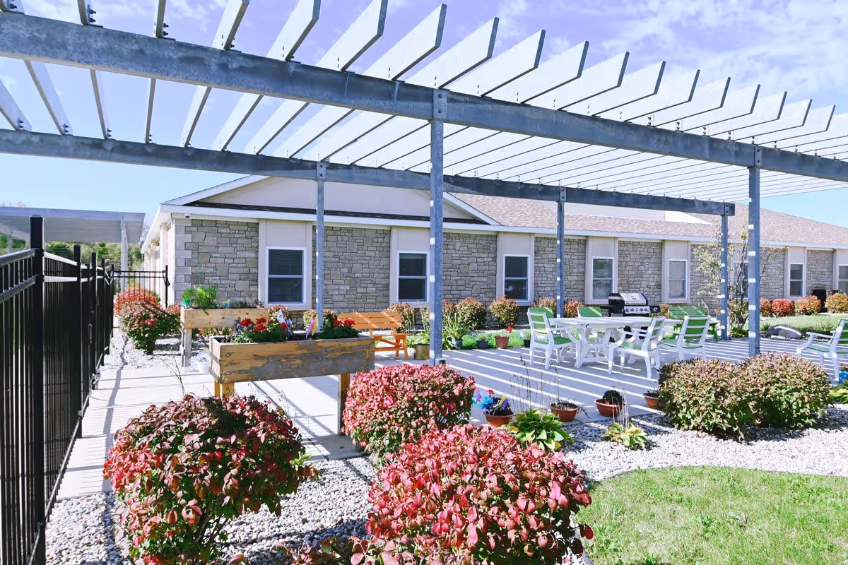 Outdoor patio area at Vista Springs Washington Place featuring a pergola, white patio table with green and white chairs, a barbecue grill, wooden planter boxes with flowers, and neatly trimmed bushes surrounded by white gravel and green grass.