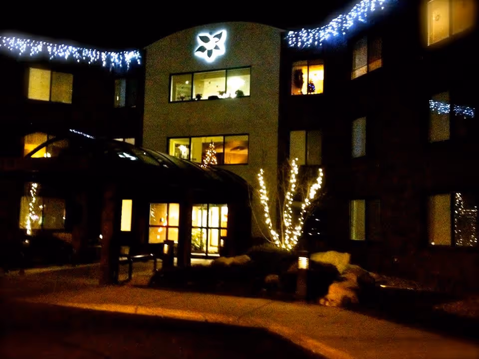 Night view of the entrance to Robbinswood Assisted Living Community building decorated with white string lights along the roofline and on a tree near the entrance. The building has multiple windows with some interior lights on, and a lit logo above the entrance.