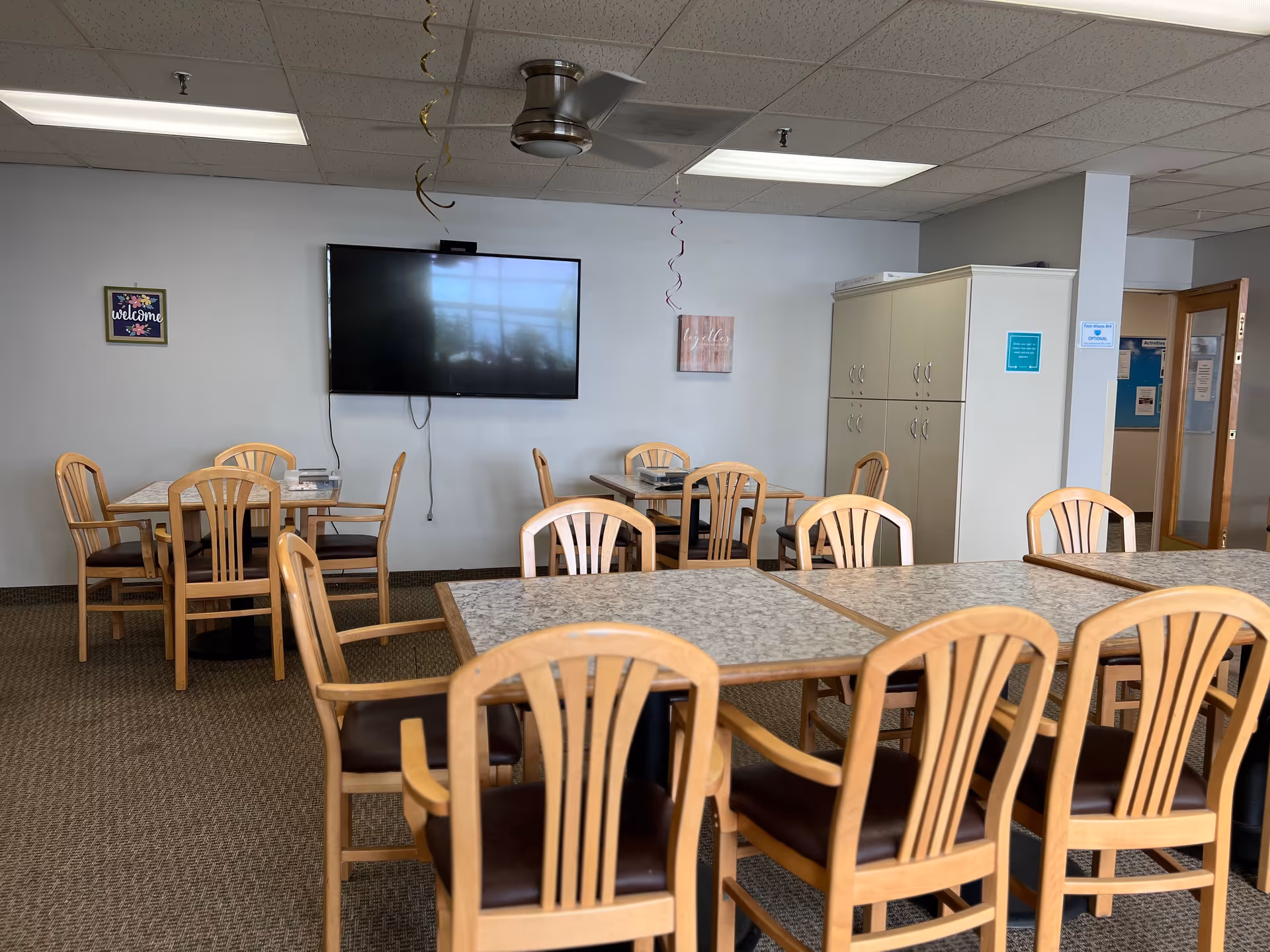 A communal dining area with multiple tables and wooden chairs with dark cushions. A large flat-screen TV is mounted on the wall, and there are decorative signs and a ceiling fan above. A cabinet and an open door leading to another room are visible in the background.