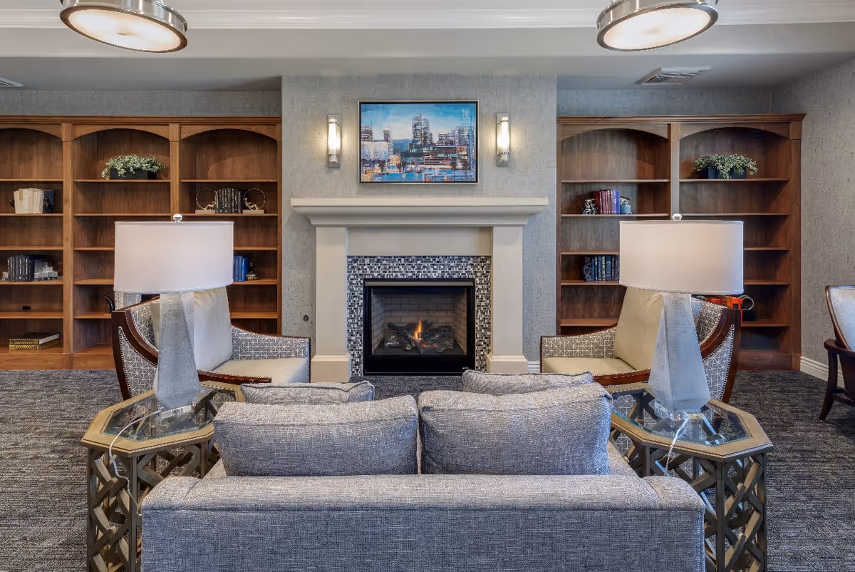A cozy living room area with a gray couch and two armchairs facing a fireplace with a mosaic tile surround. There are two side tables with lamps on either side of the couch. Behind the fireplace are built-in wooden bookshelves with some books and decorative plants. A framed cityscape painting hangs above the fireplace.