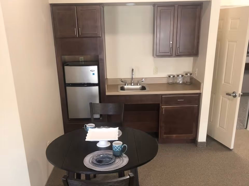 A small kitchenette area with dark wood cabinets, a stainless steel mini refrigerator, a sink, and a countertop with three glass jars. In front of the kitchenette is a round dark wood table set with two place settings, each with a plate, bowl, and mug. An open door is visible to the right.