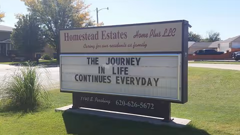 Outdoor sign for Homestead Estates Home Plus LLC on a grassy area with a street and houses in the background. The sign reads: 'THE JOURNEY IN LIFE CONTINUES EVERYDAY'.