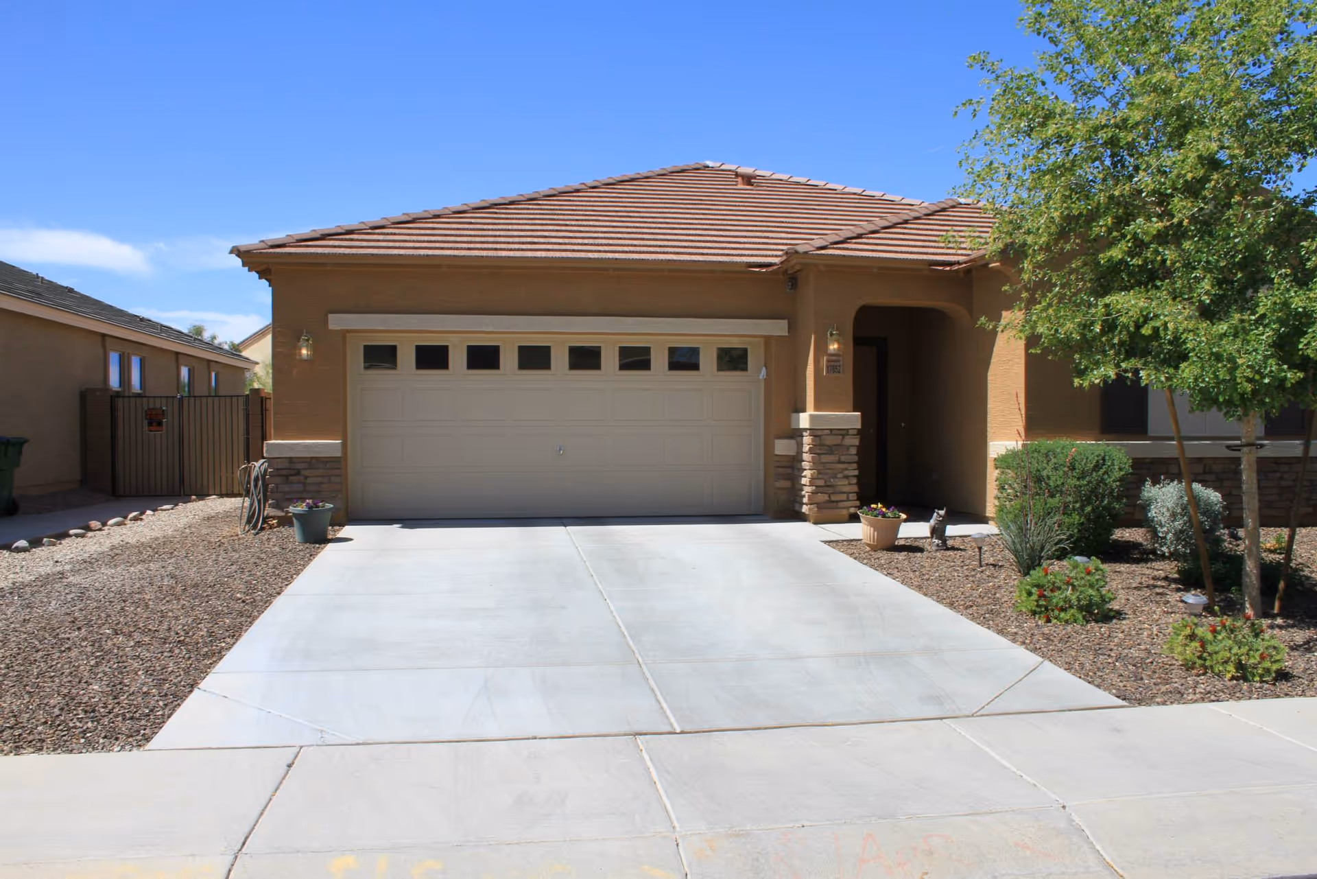 Front exterior view of a single-story house with a tiled roof, a double garage door, a concrete driveway, and desert landscaping with rocks, shrubs, and a tree.