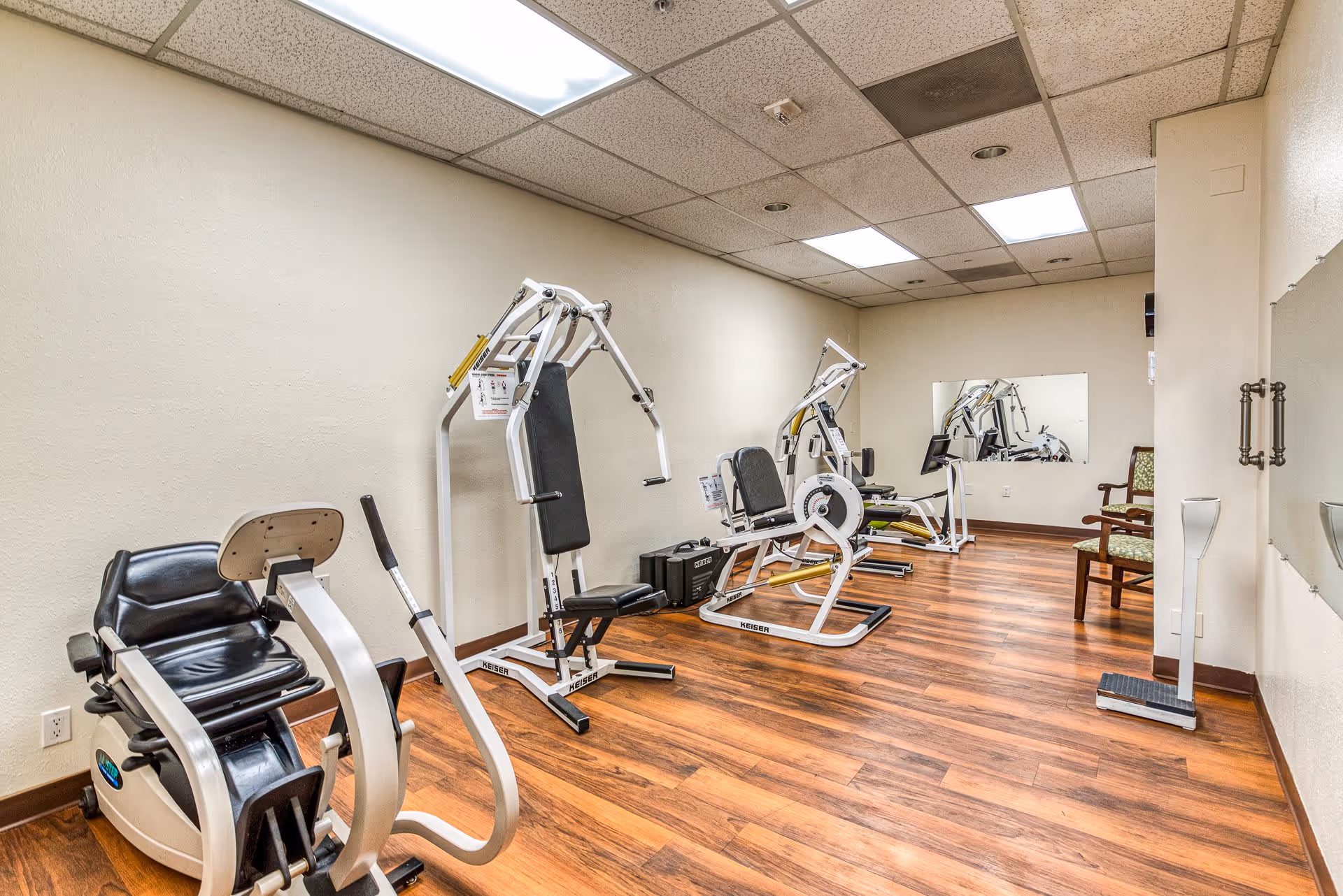 A small exercise room with wooden flooring and beige walls, equipped with several pieces of fitness equipment including a recumbent bike and weight machines. There is a large mirror on the far wall and a chair in the corner. The ceiling has recessed lighting and some ceiling tiles appear discolored.