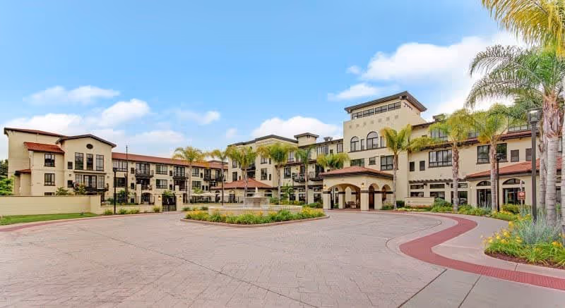 Exterior view of a large, multi-story senior living facility with beige walls, red-tiled roofs, and multiple balconies. The building is surrounded by palm trees and landscaped greenery, with a circular driveway and a fountain in front under a partly cloudy sky.