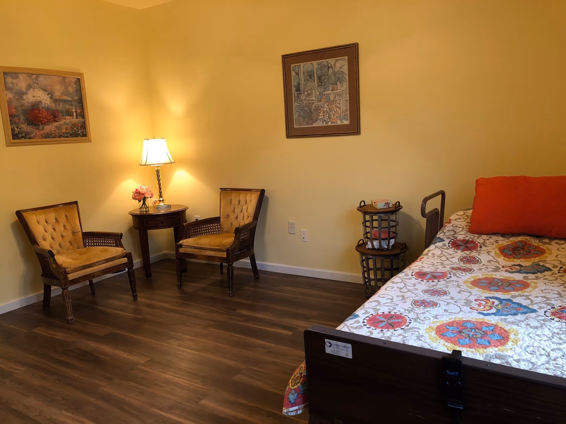 A cozy bedroom with two vintage-style armchairs and a small round wooden table with a lamp and a flower vase between them. The bed has a colorful patterned quilt and an orange pillow. Two framed paintings hang on the yellow walls, and there are two stacked baskets near the bed.
