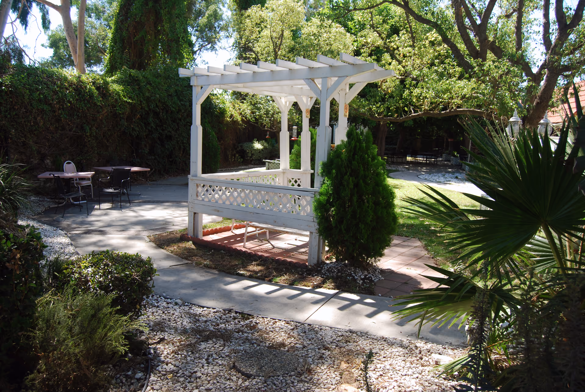 Sunlit outdoor garden patio with a white wooden pergola over a bench, surrounded by trees, shrubs, and patio tables.