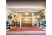 Entrance to Windsor El Camino Care Center with glass double doors, green benches on either side, potted plants hanging on the walls, and a red welcome mat on the ground.