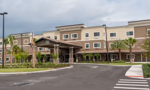 Exterior view of a modern three-story senior living facility with a covered entrance, palm trees, and a driveway under a cloudy sky.