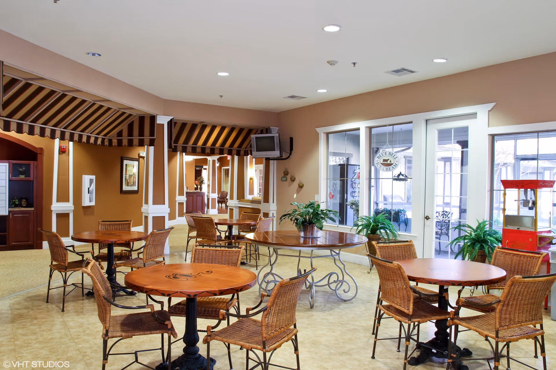 A bright and inviting common area with several round wooden tables and wicker chairs arranged around the room. There are potted plants on some tables and near the windows. The walls are painted beige, and there are large windows and glass doors letting in natural light. A small popcorn machine is visible on the right side. The ceiling has recessed lighting, and there is a small wall-mounted TV in the background.