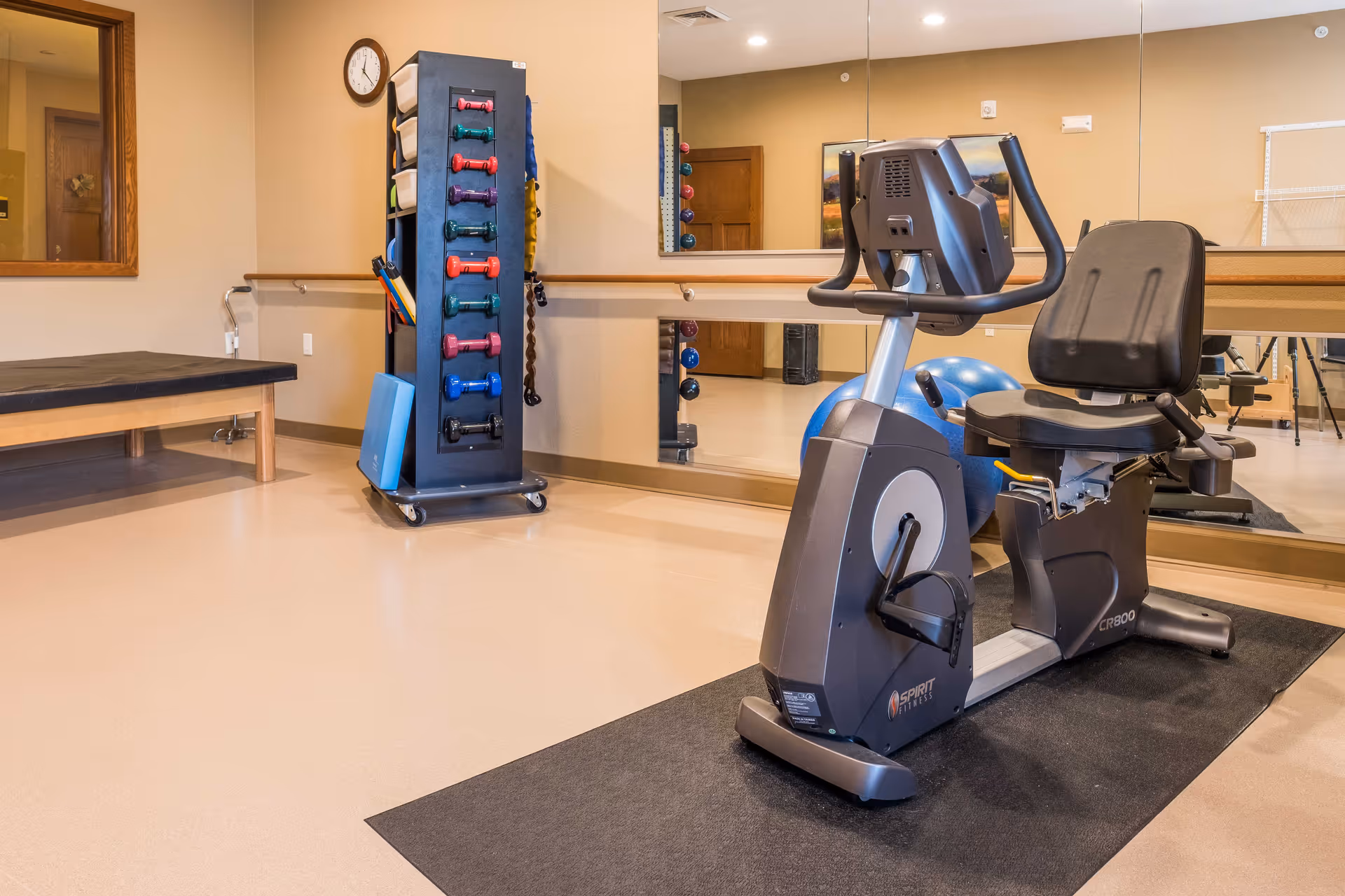 A senior living facility exercise room with a recumbent stationary bike on a black mat, a rack holding colorful dumbbells, a blue exercise ball, a padded bench, and a large wall mirror reflecting the room.