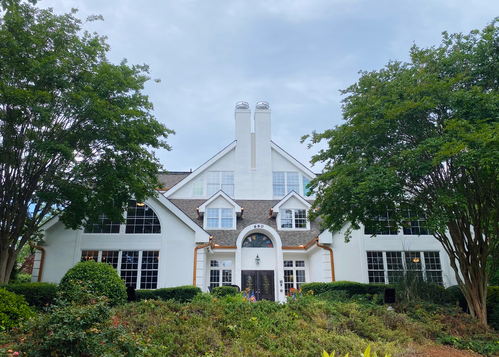 Front exterior of a white multi-story senior living building with a central arched entrance, large windows, and surrounding trees and landscaping.
