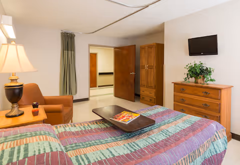 A tidy bedroom featuring a bed with a colorful quilt, an armchair, a wooden dresser with a small TV, and an open door leading to a hallway.