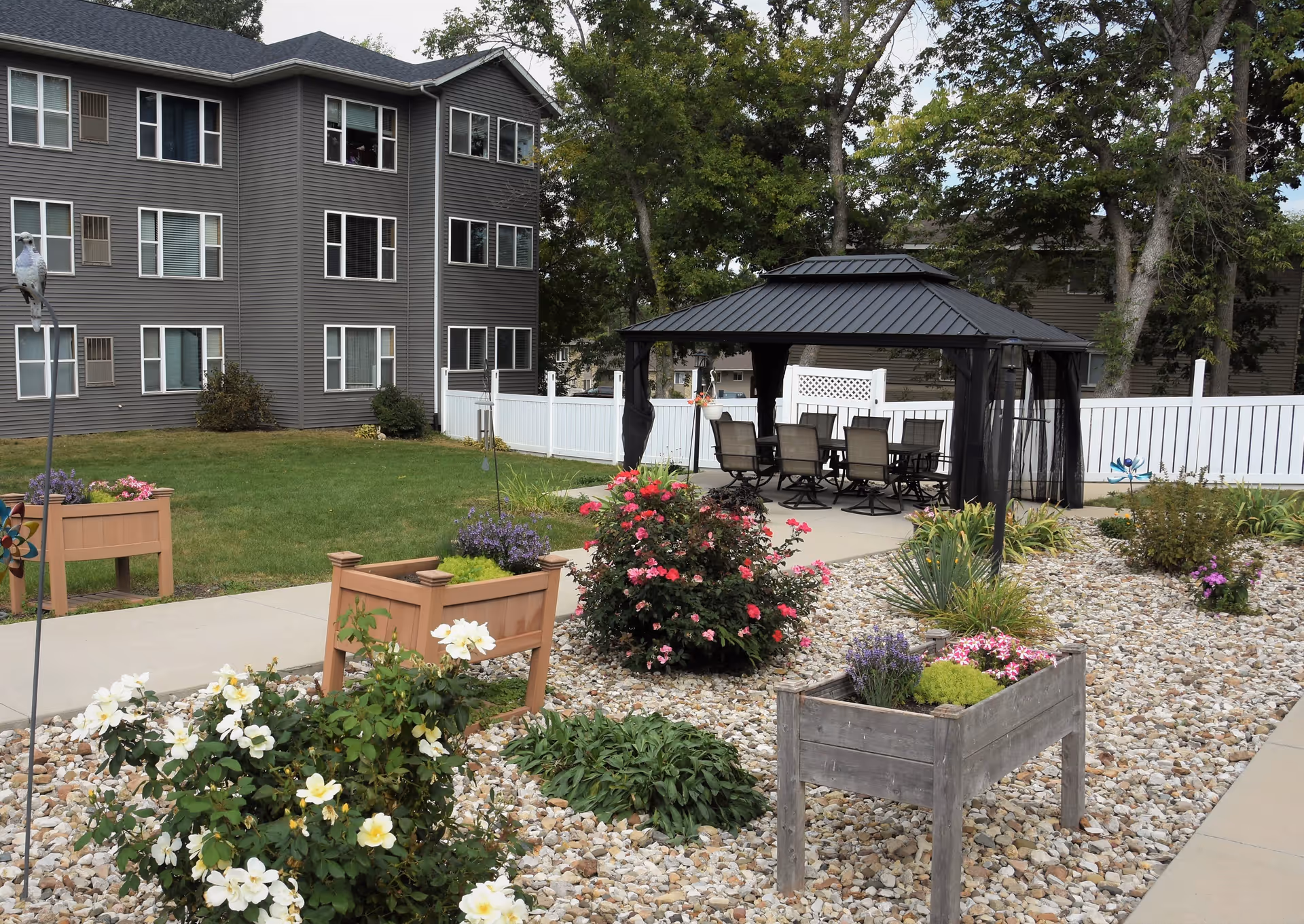 Outdoor garden area at Garnett Place featuring a gravel bed with various flowering plants and bushes, raised wooden planters, a concrete pathway, a black metal gazebo with a dining table and chairs underneath, and a multi-story gray building in the background surrounded by trees and a white fence.