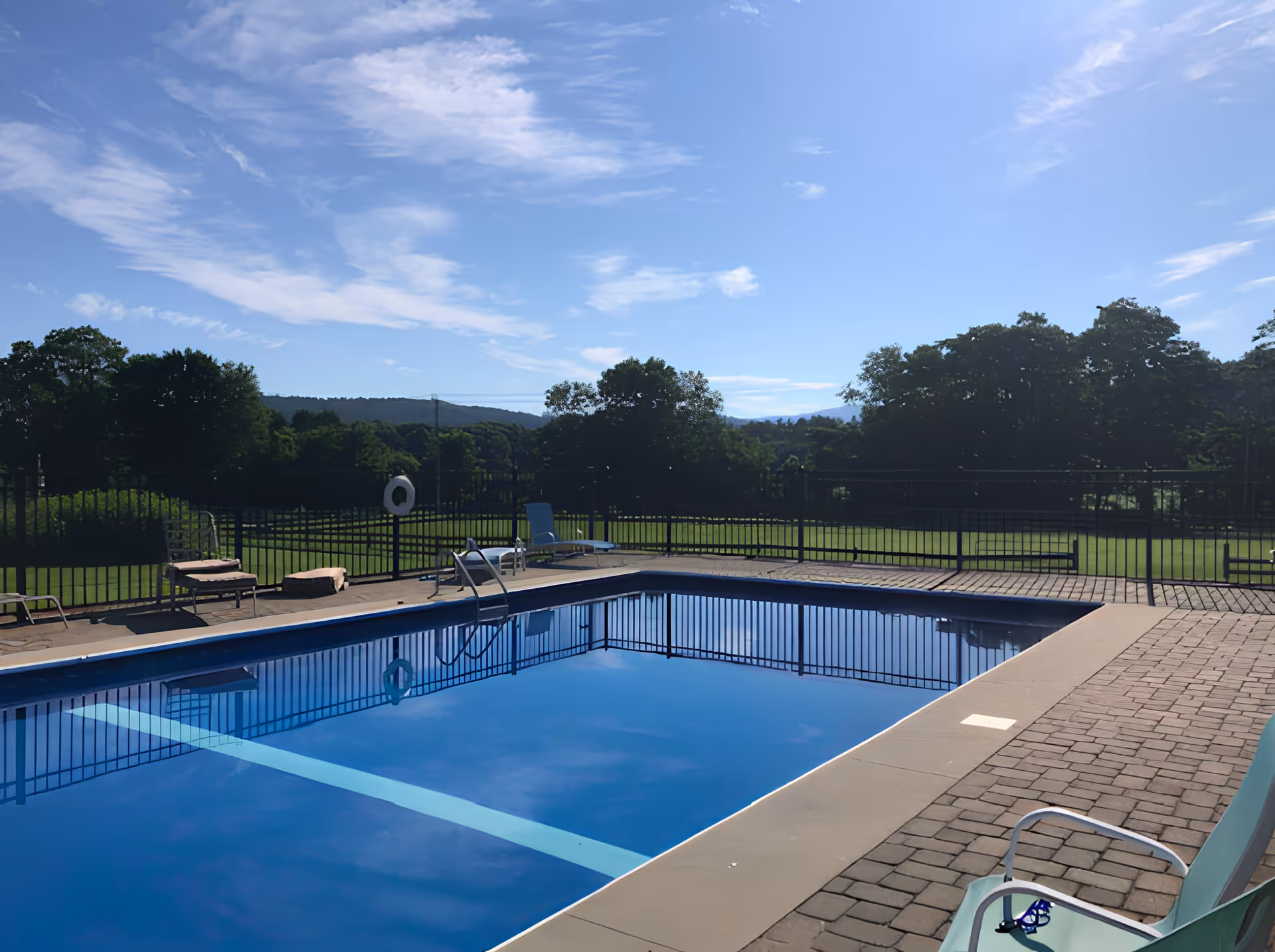 Outdoor swimming pool with clear blue water surrounded by a paved deck with lounge chairs. The pool area is fenced and beyond it is a grassy field with trees and hills under a partly cloudy blue sky.