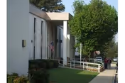 Exterior view of a white brick building with an American flag mounted near the entrance. There is a wheelchair ramp with white railings leading to the entrance, and a sidewalk running alongside the building. Trees and bushes are visible near the building and along the sidewalk.