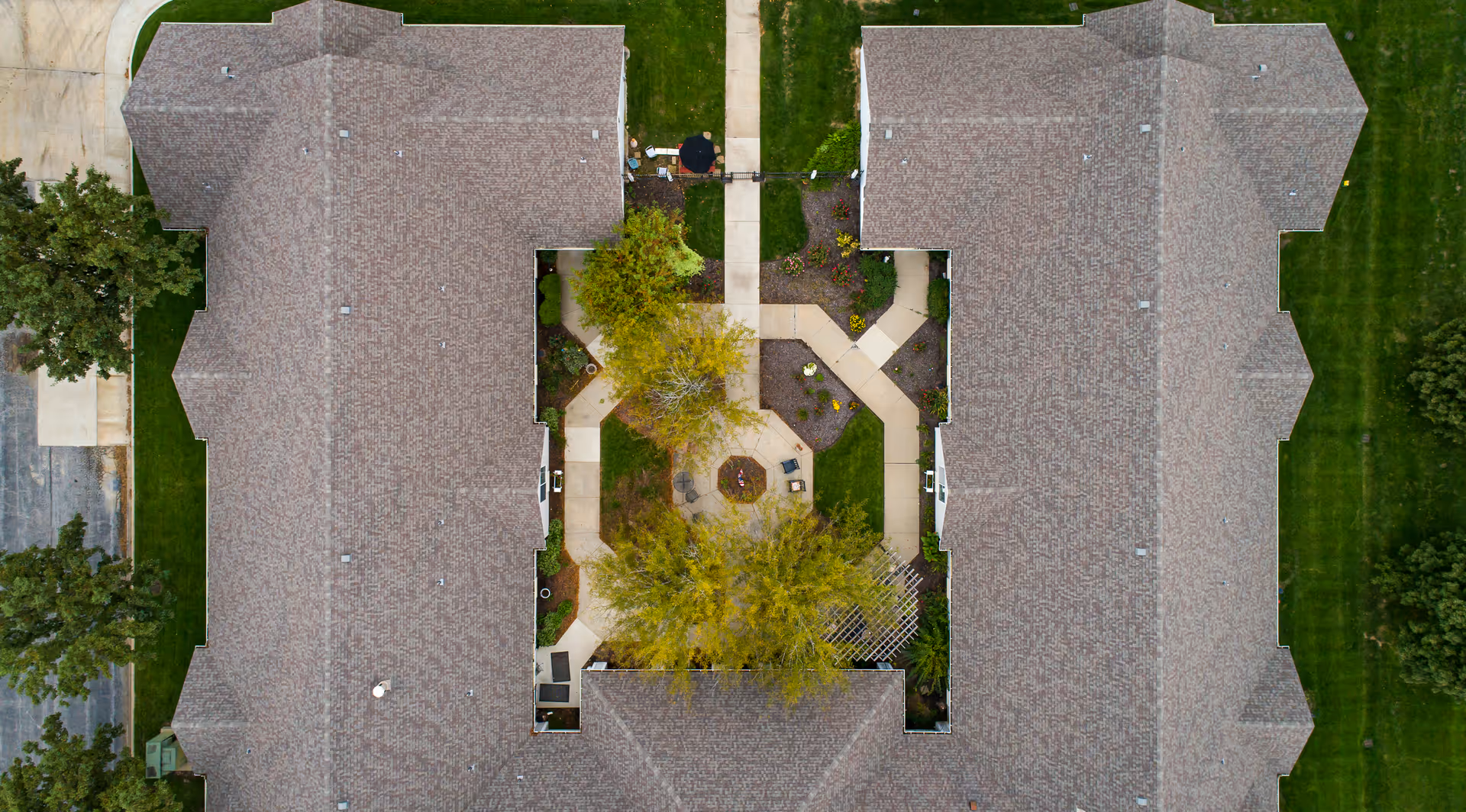 Aerial view of a senior living facility showing two large buildings with a landscaped courtyard in the center. The courtyard has paved walkways, trees, and seating areas. Surrounding the buildings are green lawns and some trees.