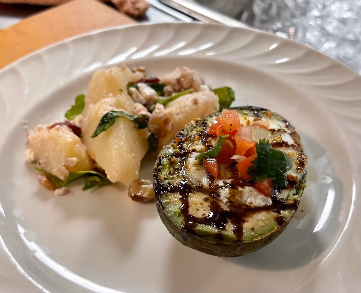 A white plate with a grilled avocado half topped with diced tomatoes, onions, and cilantro, drizzled with a dark balsamic glaze, accompanied by a side of potato salad with greens and walnuts.