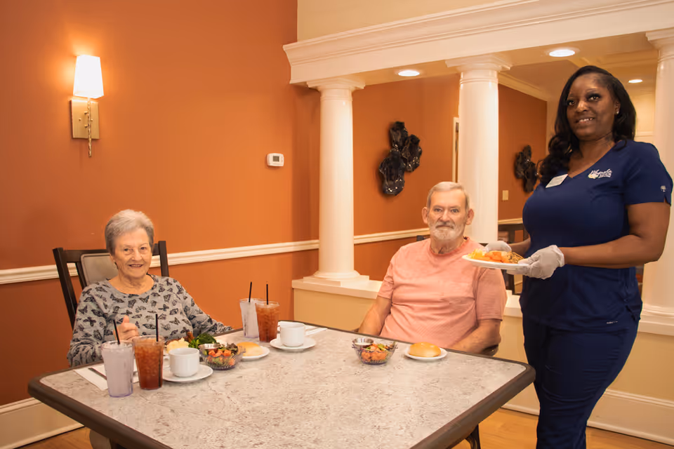 Two elderly individuals sitting at a dining table with drinks and food, while a staff member in navy scrubs stands beside them holding a plate of food in a warm, well-lit room with orange walls and white columns.