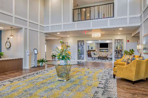 Spacious senior living facility lobby with high ceilings, a large patterned area rug, a round glass table with a floral arrangement in the center, yellow upholstered chairs on the right, and a sitting area with armchairs and a TV in the background.