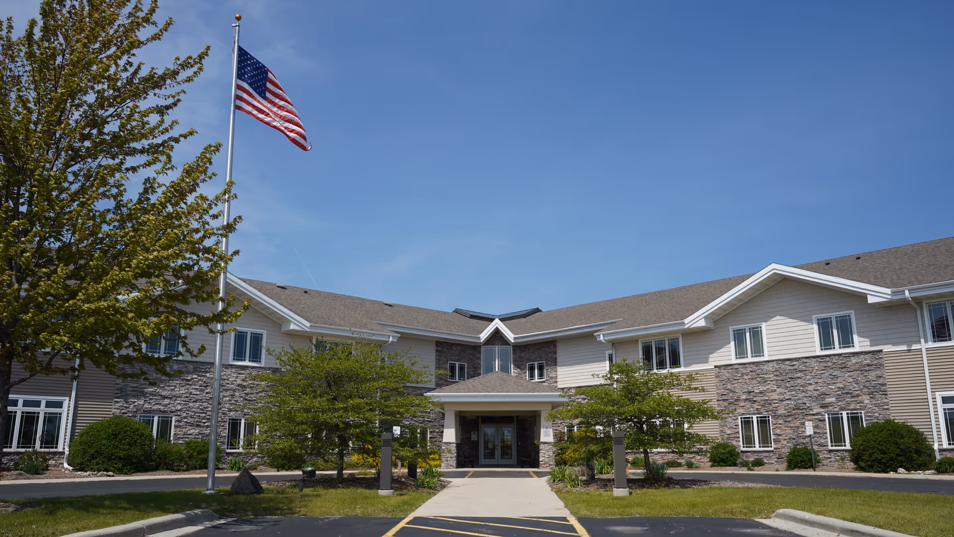 Front exterior view of a two-story senior living facility building with a stone and siding facade, a covered entrance, several windows, green trees and bushes, a flagpole with an American flag, and a clear blue sky.