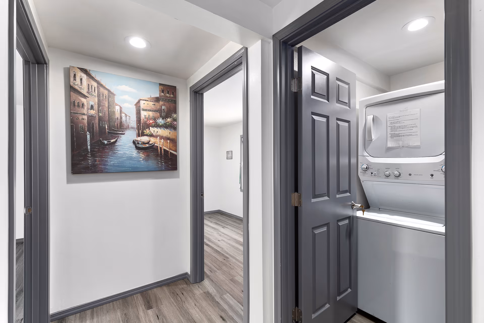 Hallway with wood-look flooring, a framed canal painting on the wall, open doorways, and a stacked washer-dryer in a closet.