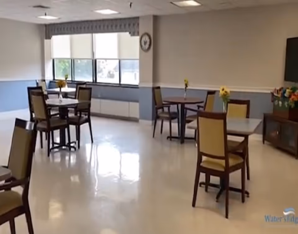 A bright dining room with several small tables and chairs arranged neatly. Each table has a small vase with yellow flowers. Large windows with blinds allow natural light to fill the room. A clock is mounted on the wall, and a cabinet with decorative flowers is visible on the right side.