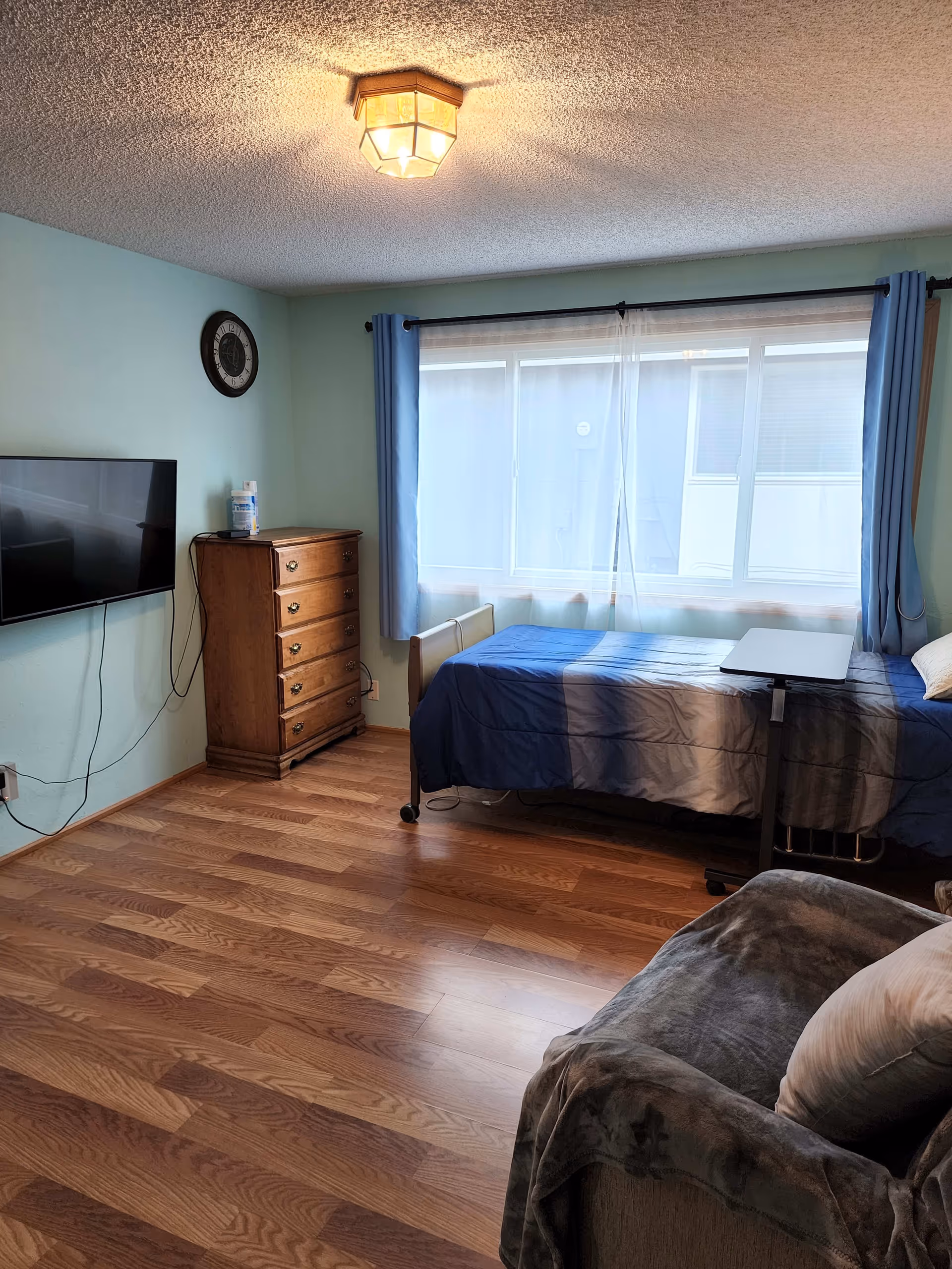 A cozy bedroom with light blue walls and wood flooring. The room features a single bed with a blue and gray gradient bedspread, a small overbed table, a wooden chest of drawers, a wall-mounted flat-screen TV, a clock on the wall, and a window with sheer white curtains and blue drapes. There is also a cushioned armchair with a gray throw and a pillow.