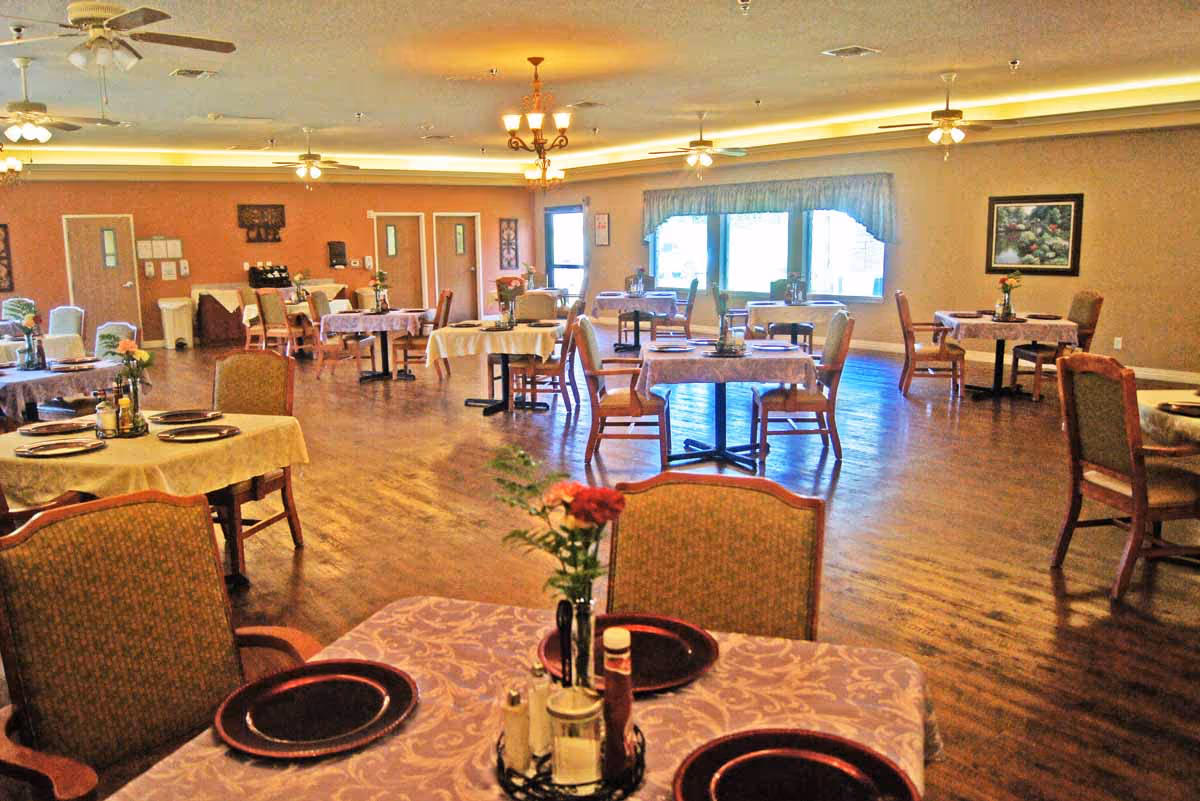 Spacious dining room with multiple round and rectangular tables set with plates and floral centerpieces and large windows along one wall.