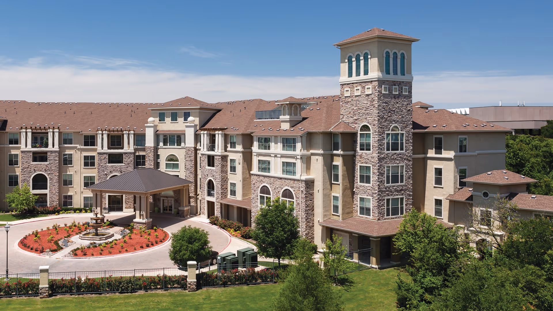 Exterior view of Atria Canyon Creek, a multi-story senior living facility with stone and beige stucco walls, a covered entrance with a circular driveway, landscaped garden with a fountain, and a tall tower-like structure on the right side under a clear blue sky.