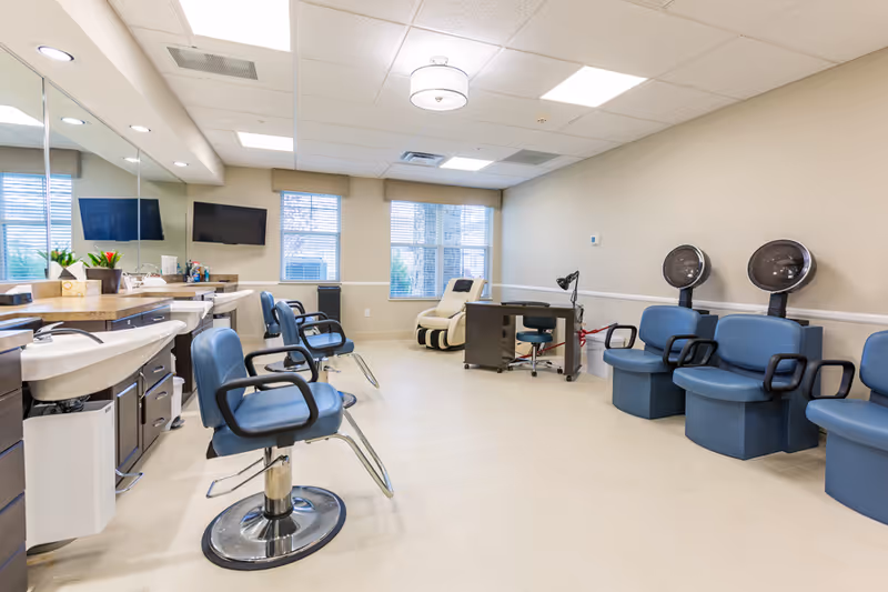 Interior of a salon area in a senior living facility with blue salon chairs, hair washing sinks, hair drying stations, a massage chair, and a manicure table under bright ceiling lights and large windows.