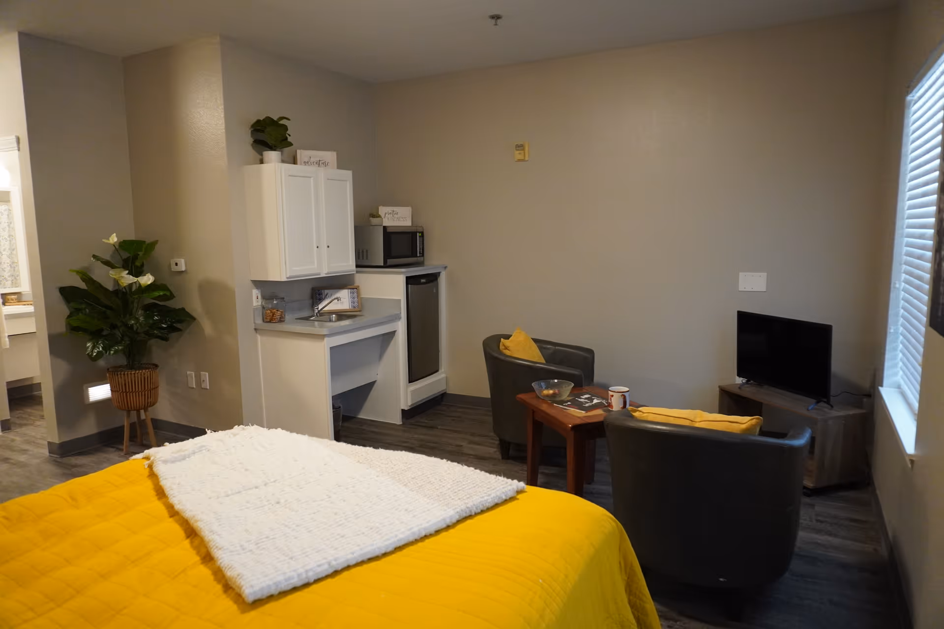 A cozy senior living room with a yellow bedspread and white blanket on the bed, two gray armchairs with yellow pillows around a small wooden table holding a bowl and a mug, a small TV on a wooden stand near a window with blinds, and a kitchenette area with a mini fridge, microwave, sink, and white cabinets. A potted plant is placed near the entrance to a bathroom.