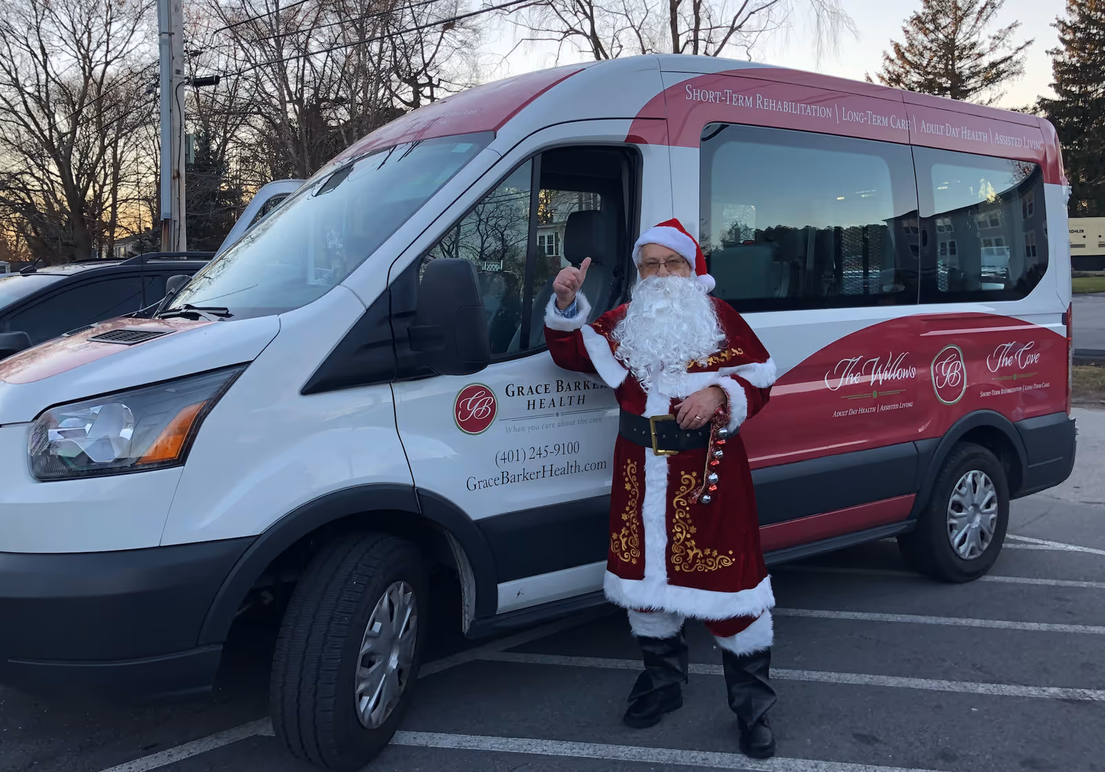 A person dressed as Santa Claus stands next to a white and red van with Grace Barker Health branding in a parking lot during dusk. The van displays contact information and services such as short-term rehabilitation, long-term care, adult day health, and assisted living.