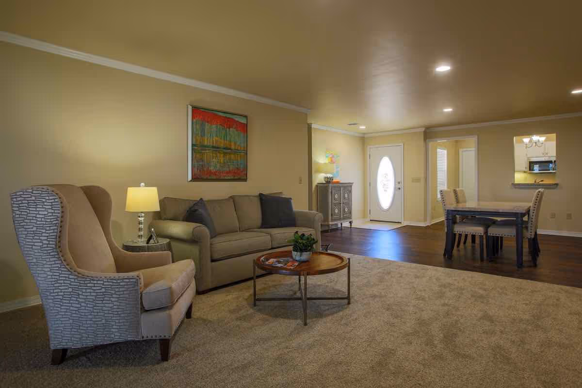 A cozy living room with a beige sofa, a patterned armchair, a round wooden coffee table with a small plant, and a side table with a lamp. The room has beige walls and carpet, with a colorful abstract painting hanging above the sofa. In the background, there is a dining area with a table and four chairs, a white front door with an oval glass window, and a small cabinet with decorative items.