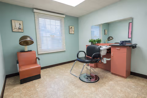 A small room with light green walls and a window with white blinds. There is a brown leather chair with a vintage hair dryer hood on the left side. On the right side, there is a black salon chair in front of a counter with a large mirror, a sink, and some folded towels. Two small framed pictures hang on the walls beside the window.